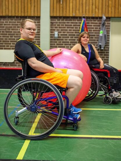 Bild från en sporthall, i förgrunden en ung man i rullstol som håller handen på en stor rosa boll. I bakgrunden en ung kvinna i rullstol.
